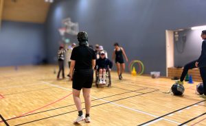 Foto på en grupp transpersoner med olika funktionsnedsättningar som utövar en parasport lek tillsammans. En person syns i förgrunden av bilden och den går med ryggen mot kameran mot resten av gruppen. Resten av gruppen är i bakgrunden och går på rad efter varandra.