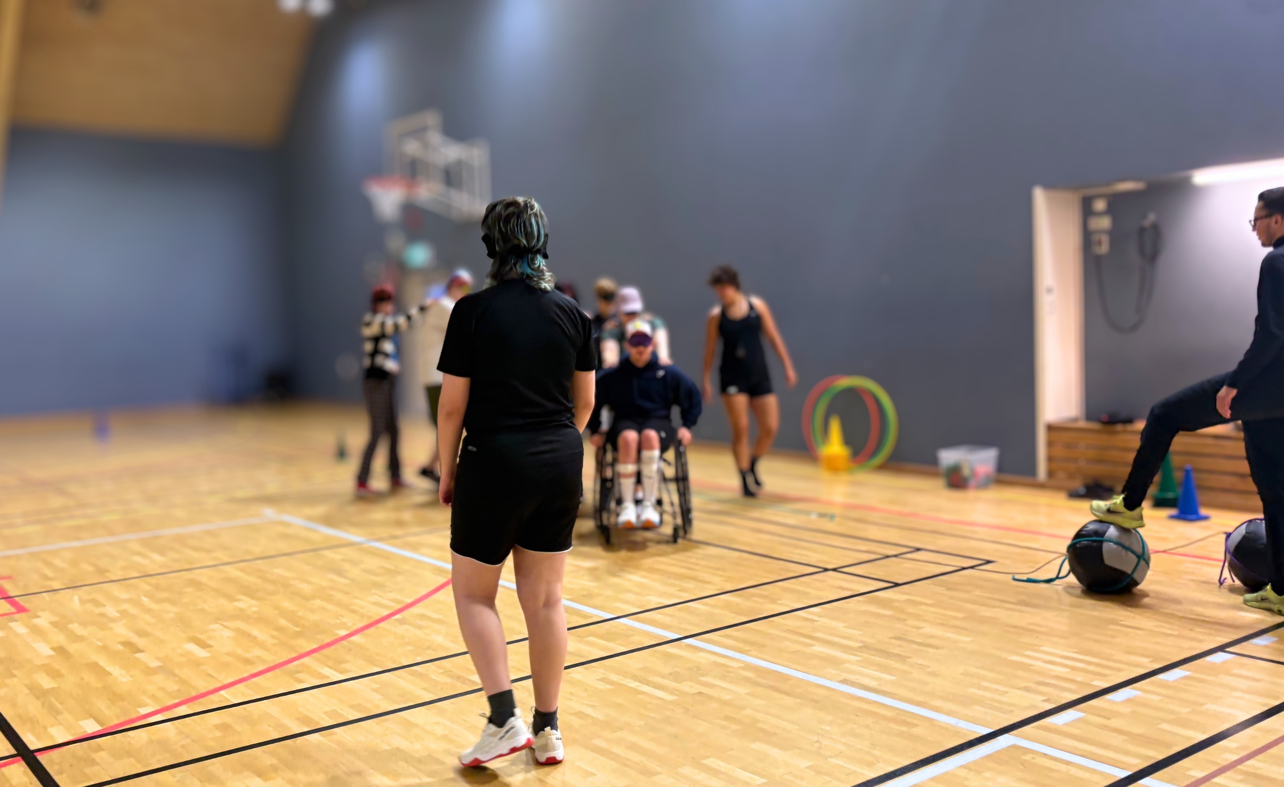 Foto på en grupp transpersoner med olika funktionsnedsättningar som utövar en parasport lek tillsammans. En person syns i förgrunden av bilden och den går med ryggen mot kameran mot resten av gruppen. Resten av gruppen är i bakgrunden och går på rad efter varandra.