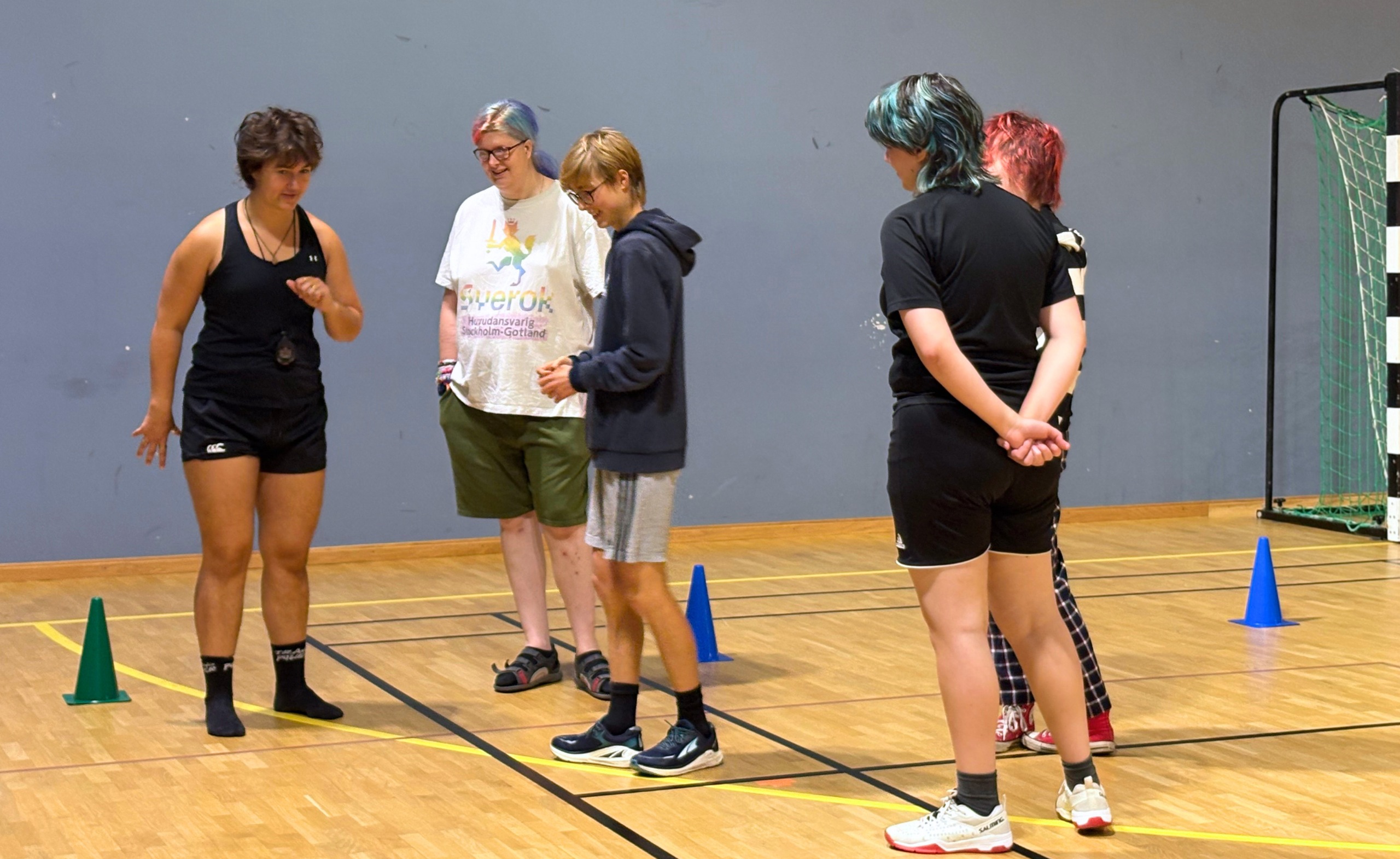 Foto på en grupp av fem transpersoner som utövar en parasport-lek tillsammans. En står och visar de andra vad de ska göra i leken. Alla ler och är glada.