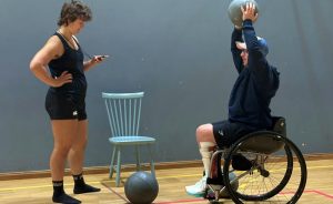 Foto på två transpersoner som utövar en parasport-lek. Den ena står upp och håller i ett tidtagarur, den andre använder rullstol och sitter och håller en boll över huvudet.